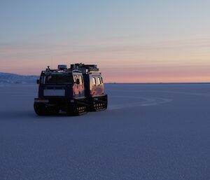 A blue Hägglunds vehicle is on sea-ice that stretches into the distance. There is an ice covered shoreline in the background to the left of frame.