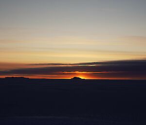 The orange glow of sunrise can be seen on the horizon against a line of clouds, across sea-ice that stretches to the horizon. There are a couple of rocky islands visible in the distance.