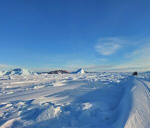 A Hägglunds vehicle can be seen in the distance travelling over snow formed into sastrugi on sea-ice. There are ice bergs and a rocky island in the far distance.