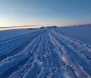 A large crack in the sea ice stretches away from the camera. It has started to freeze back over. In the distance an iceberg is frozen into the sea-ice.