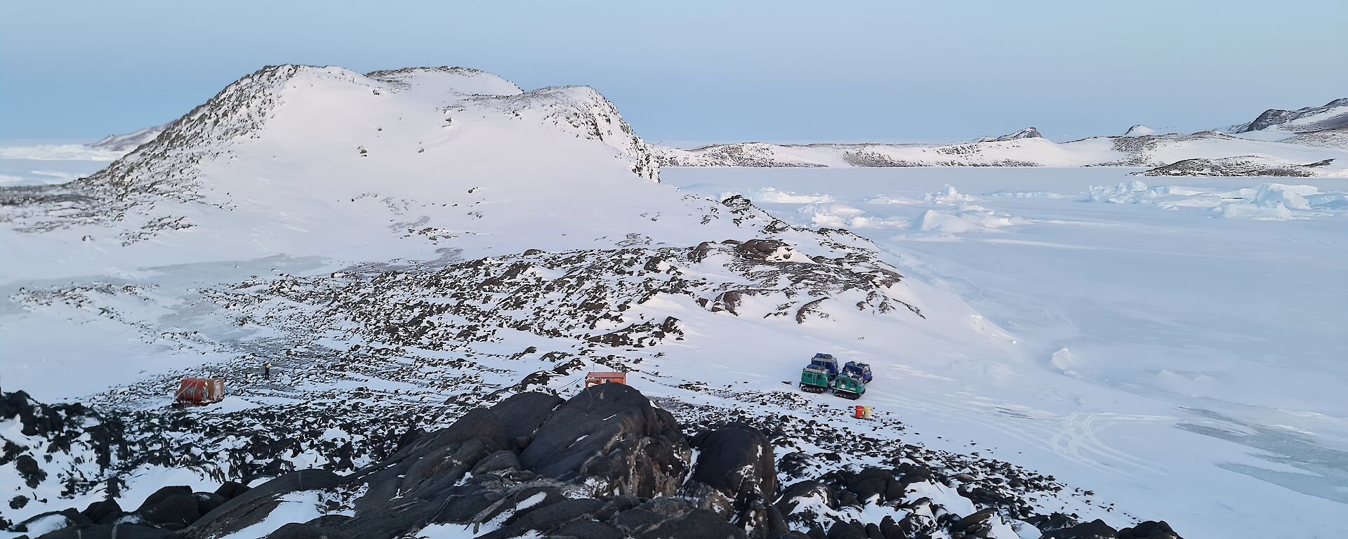 The camera is looking down on a rocky, snow covered island. Two Hägglunds vehicles are parked and there is a dark orange hut and van on the island.