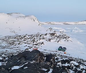 The camera is looking down on a rocky, snow covered island. Two Hägglunds vehicles are parked and there is a dark orange hut and van on the island.