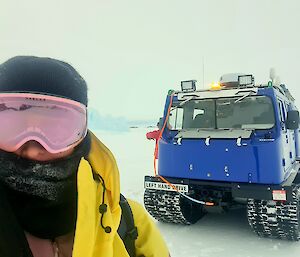 A woman wearing beanie and reflective pink goggles is looking at the camera. In the background a blue Hägglunds vehicle is parked on the ice