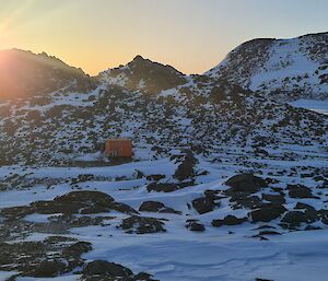 The sun is setting behind a rocky island. There are two Hägglunds vehicles parked and a dark orange hut and van on the island