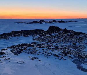 The sun is setting on the horizon. The camera is looking down from a high point over a rocky, snow covered island. There are two vehicles, a hut and a van on the middle part of the island.