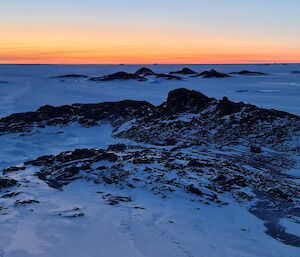 The sun is setting on the horizon. The camera is looking down from a high point over a rocky, snow covered island. There are two vehicles, a hut and a van on the middle part of the island.