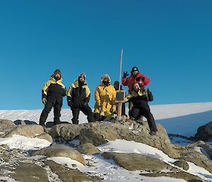 Five men and a woman are gathered around a plaque mounted on a wooden post, set into a rocky landscape. A glacier rises in the background