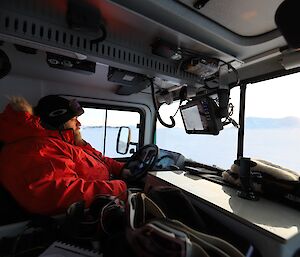 An inside photo of a Hägglunds vehicle. A man in a heavy red jacket and wearing beanie and goggles is in the drivers seat.