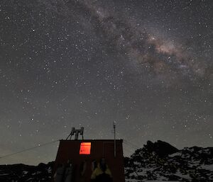 The Milky Way is visible clearly in the night sky over a hut that has a glow in the window. A woman can just be seen standing in front of the hut.