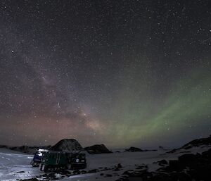 A green, orange and red aurora is visible against the night sky. There are two Hägglunds vehicles parked on the snow.