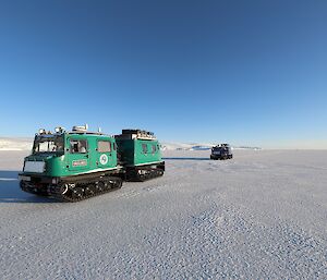 A green and a blue Hägglunds vehicle are on the sea ice on a brightly lit day. In the distance an ice plateau can be seen
