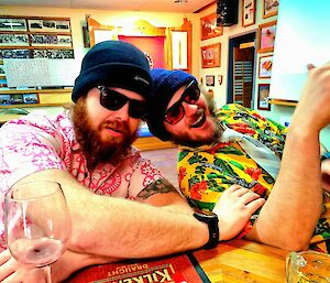 Two men are at the bar. They are wearing brightly coloured shirts, beanies, and dark sunglasses and smiling at the camera.