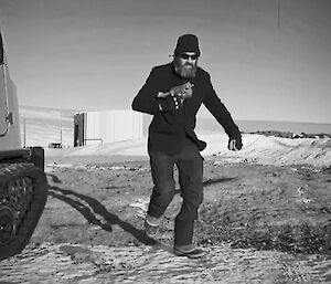 A black and white photo of a man walking towards the camera in a dark jacket, beanie, and wearing dark glasses. There is a Hägglunds vehicle parked to the left of frame.