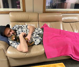 A man covered in a hot pink blanket lies on a couch cuddling a stuffed toy dog.