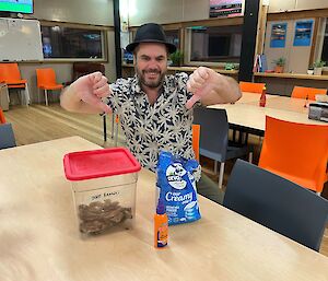 A smiling man sits at a table giving the thumbs down to a box of dried bananas, bottle of orange oil, and a packet of powdered milk.