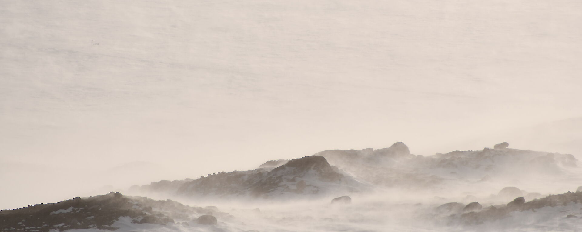 Snow blowing over some low rocky hills, blending into a grey sky in the background.