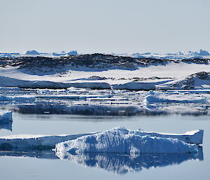 Long, low icebergs are scattered around a flat calm stretch of water. There are low rock hills in the background and the sky is clear of cloud.