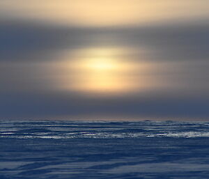 The sun is heavily filtered through some hazy cloud, and is lighting up an ice covered ground.
