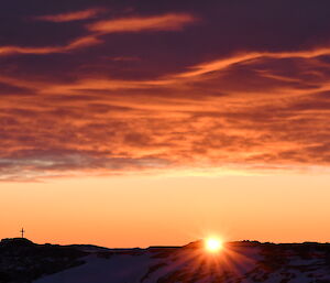 The sun is just above the horizon of silhouetted icebergs. The sky is filled with ribbons of cloud that are reflecting the colour from the sun. To the far left of the photo stands a single cross, in memory of a Casey Expeditioner who died here in 1979.