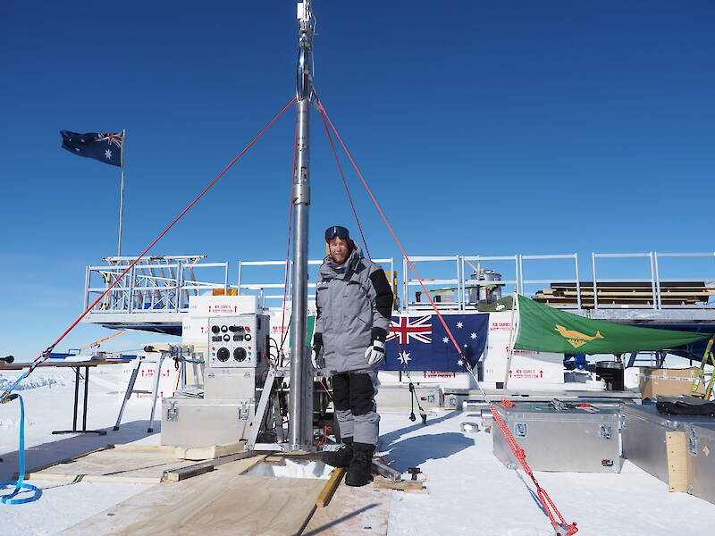 A man in warm clothing stands beside a tall metal tube. The Australian flag is flying from a raised platform behind.