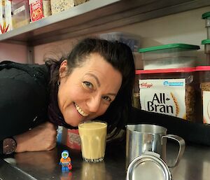 A lady is leaning over a stainless steel bench and smiling at the camera with a coffee placed on the bench in front of her. There is shelving above containing various cereals.