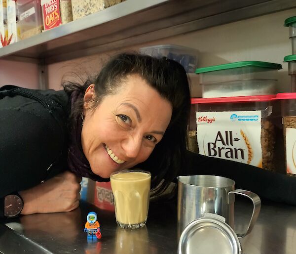 A lady is leaning over a stainless steel bench and smiling at the camera with a coffee placed on the bench in front of her. There is shelving above containing various cereals.