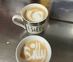Two cups filled with coffee sit on a stainless steel bench. The camera is looking down into the coffees and the name Billy is written in one cup with milk foam.