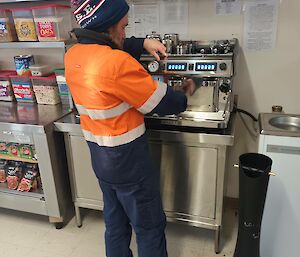 A man in bright orange hi-vis workwear is standing with his back to the camera and making a coffee at a large coffee machine. There are rows of shelves behind him with various cereals.