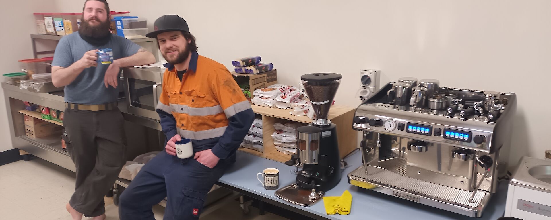 A large stainless steel coffee machine on a long table fills the right of the photo. To the left are two men in workwear, reclining against the table and an adjacent bench. They are smiling at the camera and enjoying a coffee.