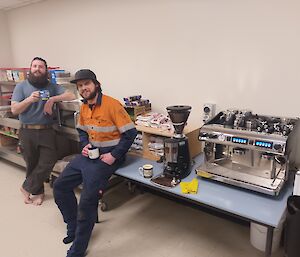 A large stainless steel coffee machine on a long table fills the right of the photo. To the left are two men in workwear, reclining against the table and an adjacent bench. They are smiling at the camera and enjoying a coffee.