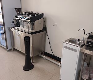 A large coffee machine sits on a stainless steel cupboard to the left of the picture. To the right of the picture is a drinking water fountain. In between the objects is some exposed pipework that runs along the wall at floor level.