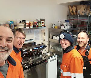 Four men in bright orange hi-vis are smiling at the camera, two on each side of the photo. In the centre of the photo is a large coffee machine, with a shelf above it containing various condiments.
