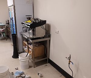 To the left of the photo is a large fridge, sited against the wall. In front of the fridge is a coffee machine that is also against the wall. There are various tools on the floor, and work is in progress on some exposed plumbing that protrudes from the floor.