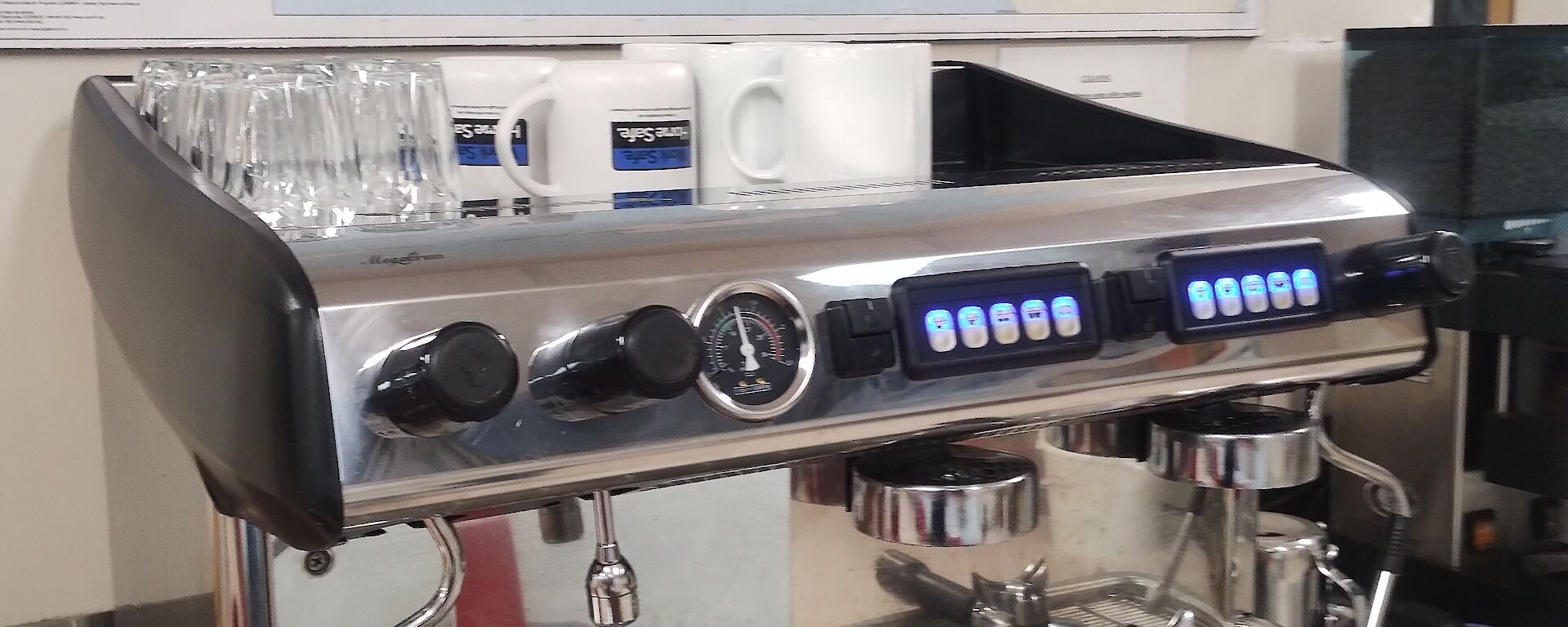 A brightly polished silver espresso coffee machine is on a stainless steel bench. On the wall above the machine is a map of Antarctica.