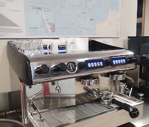 A brightly polished silver espresso coffee machine is on a stainless steel bench. On the wall above the machine is a map of Antarctica.