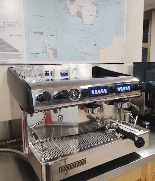 A brightly polished silver espresso coffee machine is on a stainless steel bench. On the wall above the machine is a map of Antarctica.