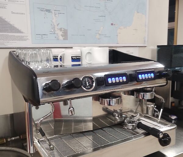 A brightly polished silver espresso coffee machine is on a stainless steel bench. On the wall above the machine is a map of Antarctica.