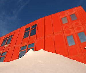 A large red metal clad building with highly reflective windows stands against the blue sky. There is a large snow drift against the building.