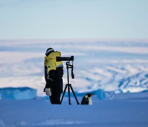 A woman is standing looking away from the camera. There is a tripod with a camera mounted and an emperor penguin looking in the opposite direction.