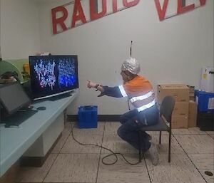 A man is sitting in a chair wearing a helmet covered in tin foil. He is pointing at a computer screen which shows a garbled picture