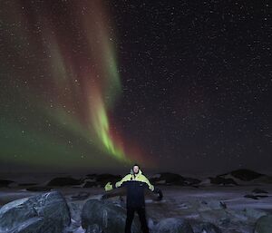 A man is looking at the camera. In the night sky behind him is a bright red, yellow and green aurora