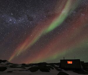 A hut with a red glow visible through the window is silhouetted by the night sky. A bright red, orange and green aurora is visible over the milky way.