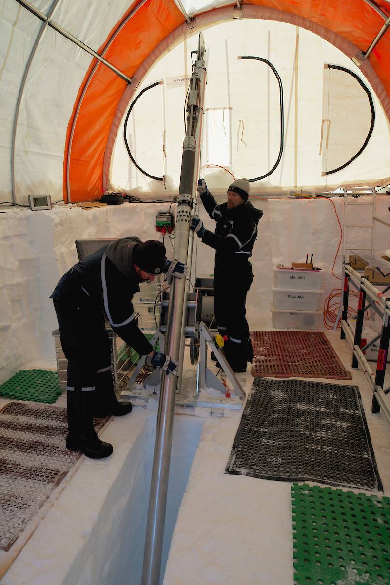 Two people inside an ice core drill tent working an ice drill with a long, steel barrel.