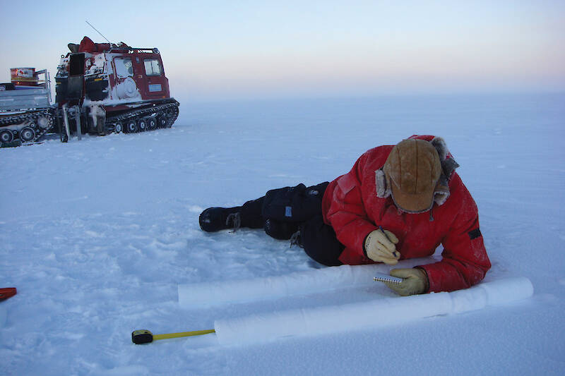 A man lying beside two ice cores in the snow, taking notes.