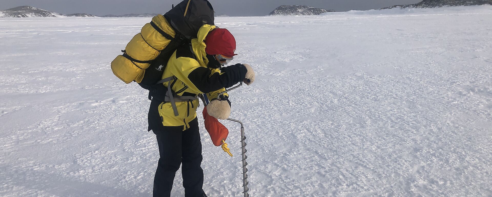 A person in yellow cold weather clothing, and carrying a backpack, is hunched over a long metal drill that is being used to bore into the ice. They are standing on a flat expanse of ice, with a gloomy overcast sky in the distant background.