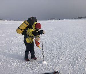A person in yellow cold weather clothing, and carrying a backpack, is hunched over a long metal drill that is being used to bore into the ice. They are standing on a flat expanse of ice, with a gloomy overcast sky in the distant background.