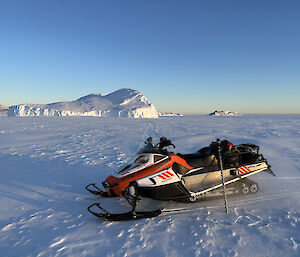 A snowmobile is parked on a flat expanse of snow covered sea ice. In the distant background is a large iceberg that has grounded on the shoreline and been surrounded by the sea ice.