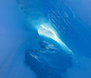 A photo from inside a cave formed by blue ice. There is a hole at the end of the cave where the light is shining through.