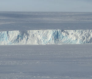 A large wall of ice stretches across the photo. In the foreground is a flat expanse of sea ice, and a snow covered ice hill rises to the rear of the picture.
