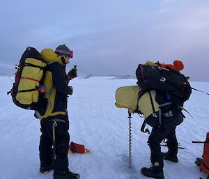Two men in yellow cold weather clothing, and carrying backpacks, are hunched over a long metal drill that is being used to bore into the ice. They are standing on a flat expanse of ice, with a gloomy overcast sky in the distant background.